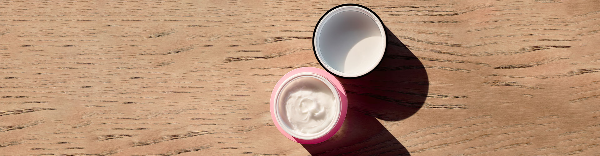 Open jar of pale pink face cream, revealing a creamy, smooth texture, placed on a light wood bathed in light.