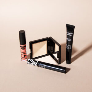 1944 Paris make-up products with compact powder, mascara, lip gloss and highlighter on a beige background.