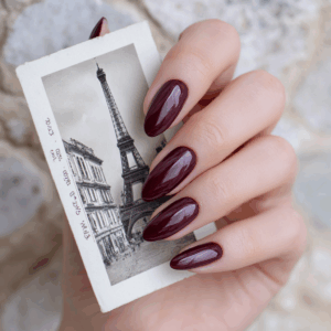 Main féminine avec vernis semi-permanent bordeaux intense 1944 Paris, manucure élégante devant une carte postale de Paris