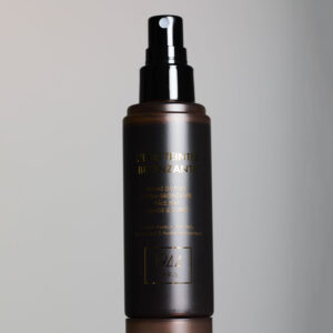 Sprayflaska av brunt glas med Eau Teintée Bronzante 1944 Paris, mot ljus bakgrund, med svart pump och guldfärgad etikett.