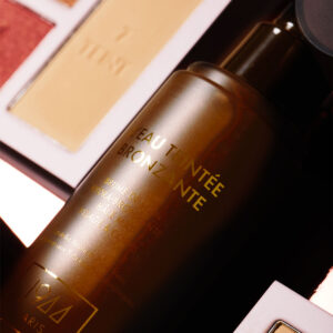 Gros plan sur le flacon d’Eau Teintée Bronzante 1944 Paris avec reflets dorés, accompagné d’une palette teint en arrière-plan flou.