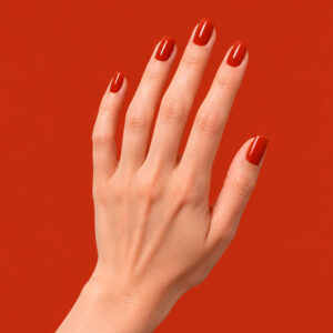 A hand with fair skin wearing Maella’s orange-red vegan semi-permanent nail varnish, against a brick-red background
